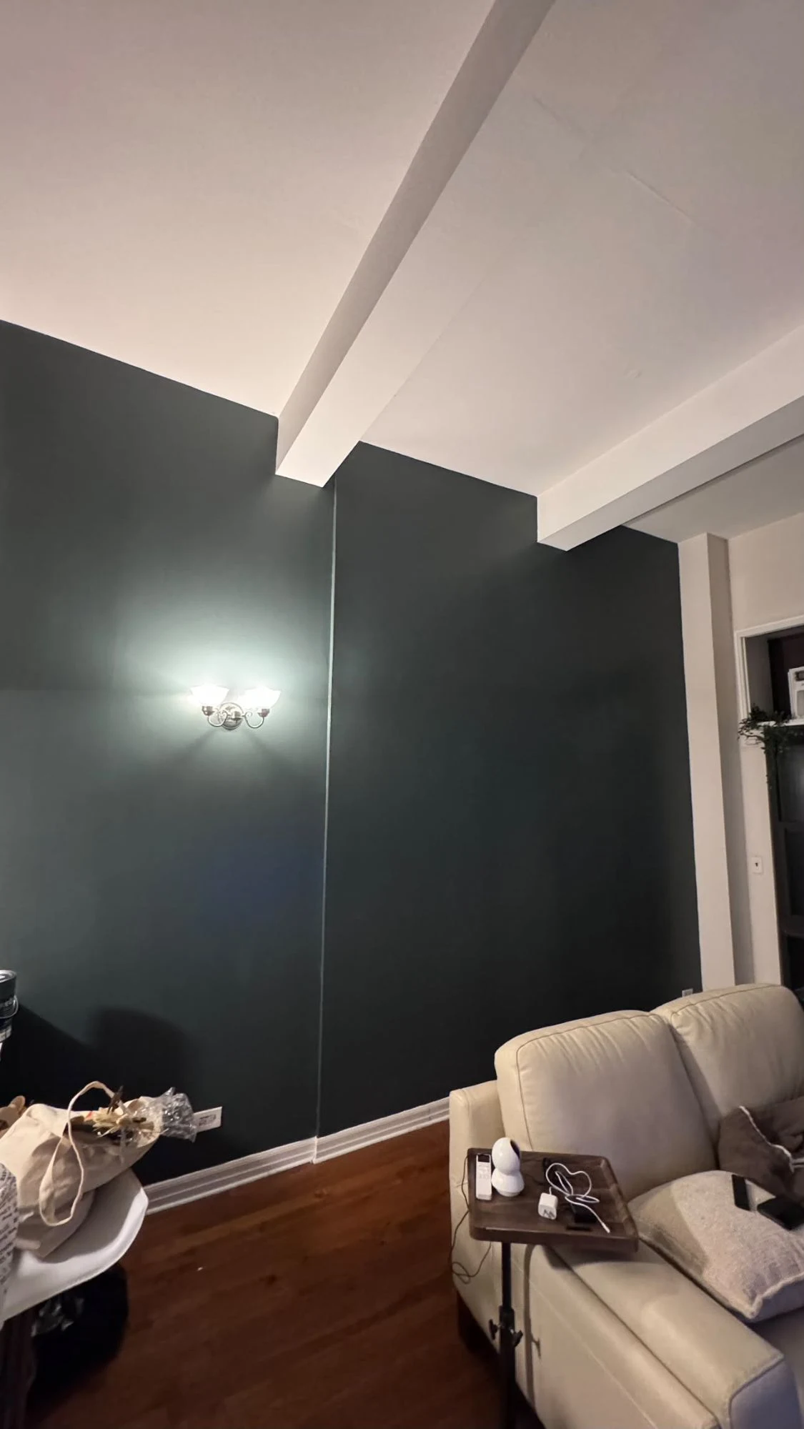 Tall apartment living room with a deep teal accent wall, a wall sconce, white ceiling beams and trim, a cream leather sofa and dark hardwood floors