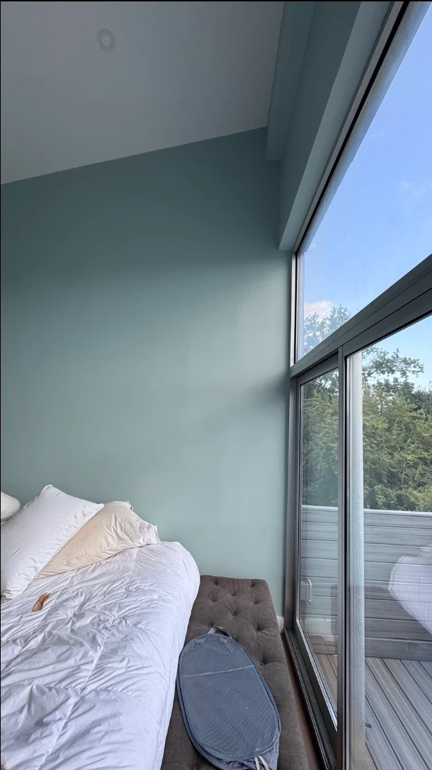 Bedroom wall in soft aqua beside a tall corner window with a balcony and tree view, and a bed with white bedding
