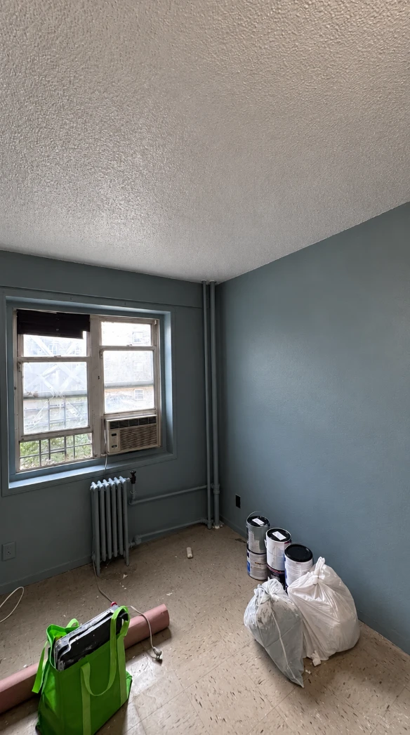 A different bedroom in another apartment, painted in deep blue-gray with matching window trim and pipes and a fresh white textured ceiling