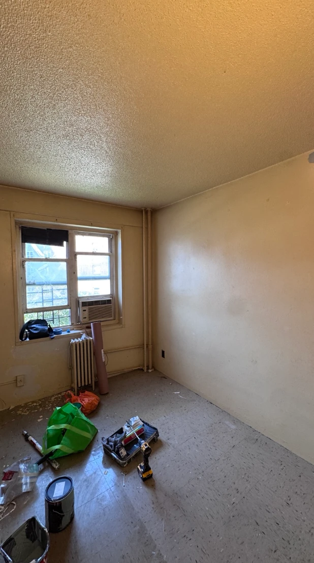 The same blue-gray bedroom before painting, with yellowed cream walls, a textured ceiling, a radiator and exposed pipes