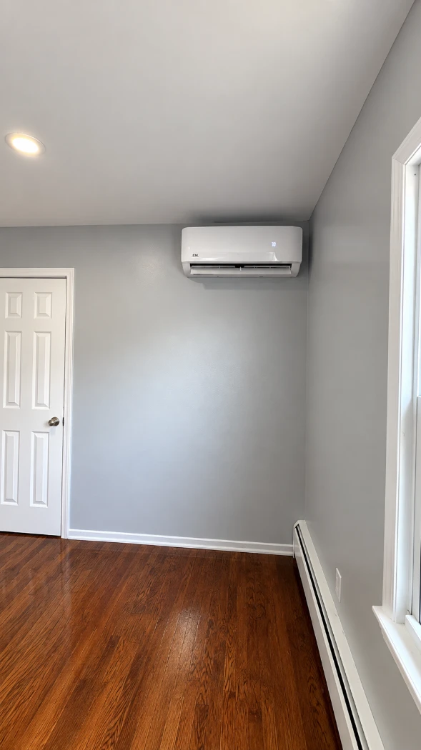Apartment bedroom with smooth cool gray walls, a white six-panel door, a wall-mounted air conditioner, white baseboards and a baseboard heater, and glossy hardwood floors