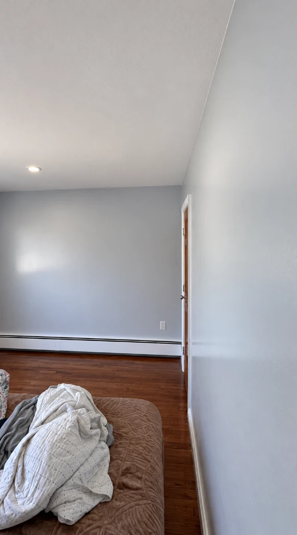 Long smooth light gray wall in an apartment bedroom with a white baseboard heater, a door with white trim and a bed in the foreground