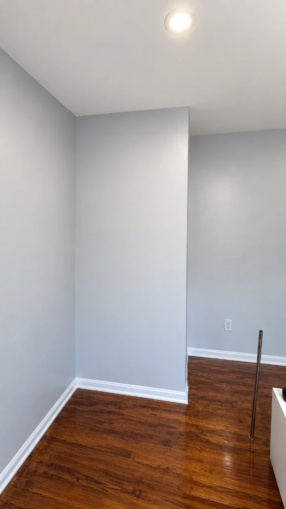 Apartment bedroom corner with smooth light gray walls meeting at sharp, straight lines, white baseboards, a recessed ceiling light and shiny hardwood floors
