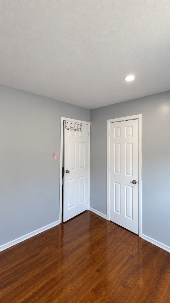 Corner of an apartment bedroom with even light gray walls, two white six-panel doors, crisp white baseboards and a recessed ceiling light above glossy hardwood floors