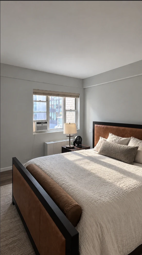 Apartment bedroom with soft gray walls and ceiling, a window with a roman shade, and a made bed with cream bedding and a leather headboard in natural light
