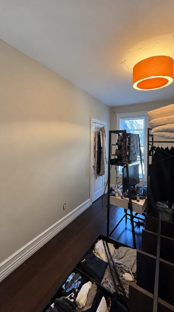 Apartment bedroom with smooth warm taupe walls, white baseboard, dark hardwood floors, a tall window and an orange pendant light