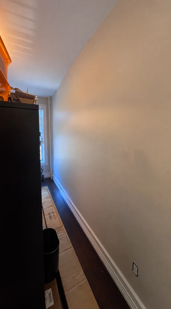 Long warm taupe bedroom wall with a white baseboard and dark hardwood floors, with a tall window at the far end