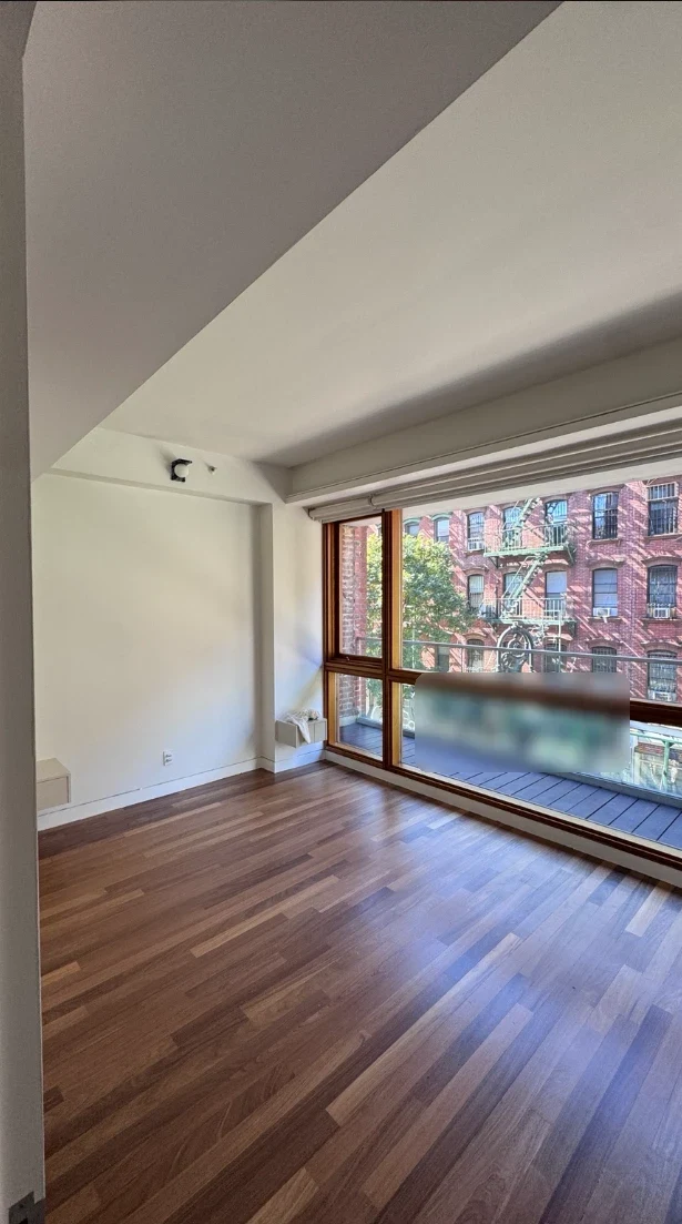 Bright apartment bedroom with smooth warm white walls and ceiling, hardwood floors and large wood-framed windows that look out to a brick building across the street