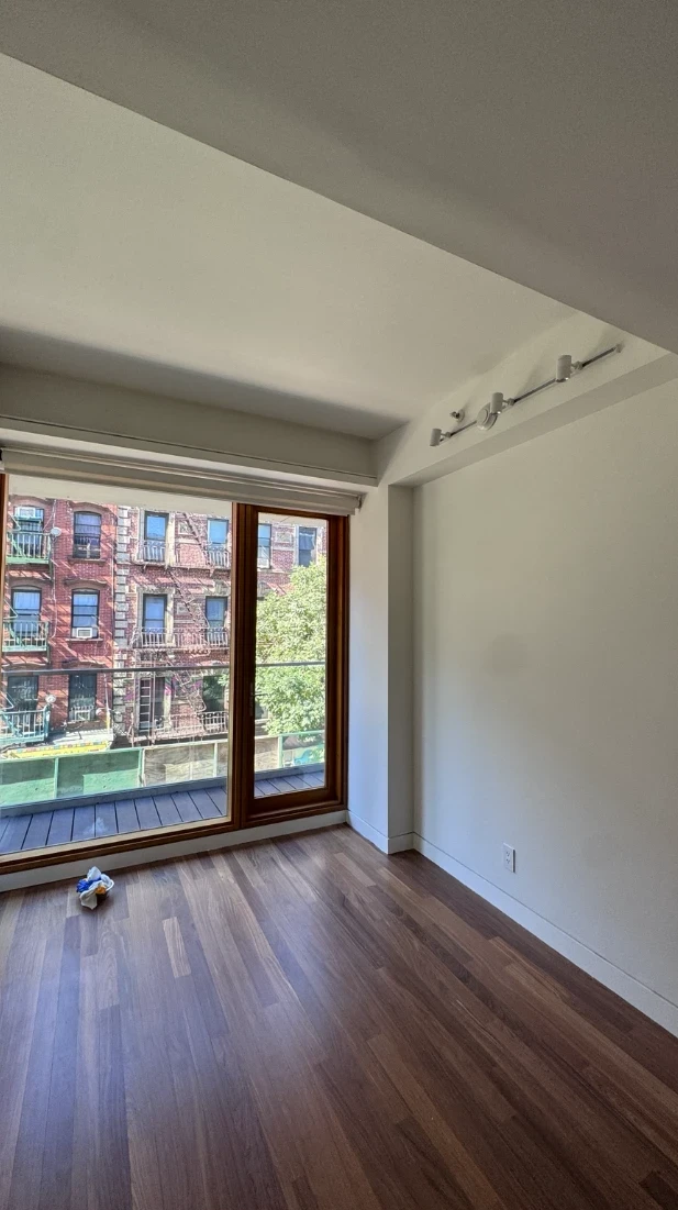 Bright apartment bedroom with smooth soft white walls and ceiling, a ceiling track light, white baseboards, hardwood floors and a wood-framed window that looks out to a brick building