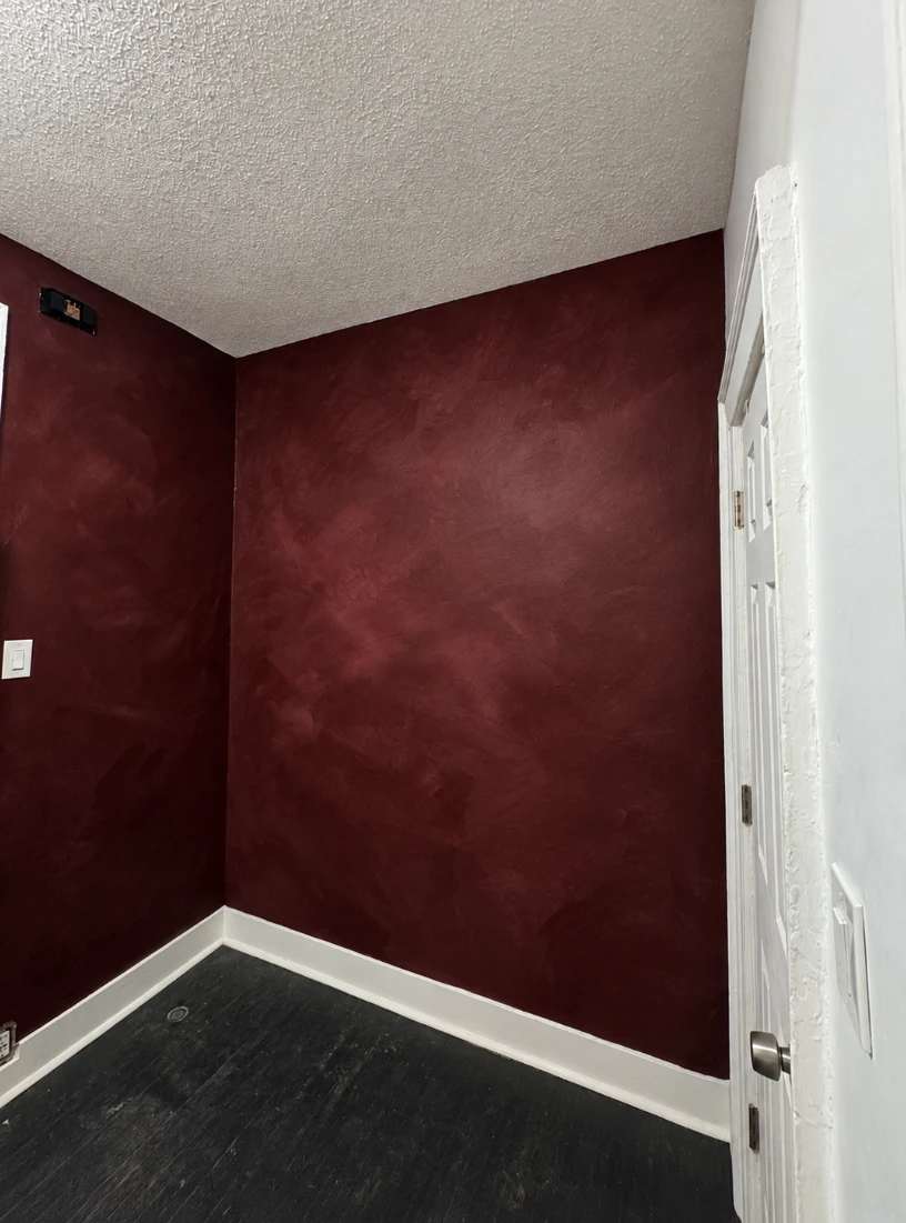 Corner of an apartment room with deep burgundy lime wash walls showing a soft, cloudy brushed texture, white baseboards, a textured white ceiling and a dark wood floor