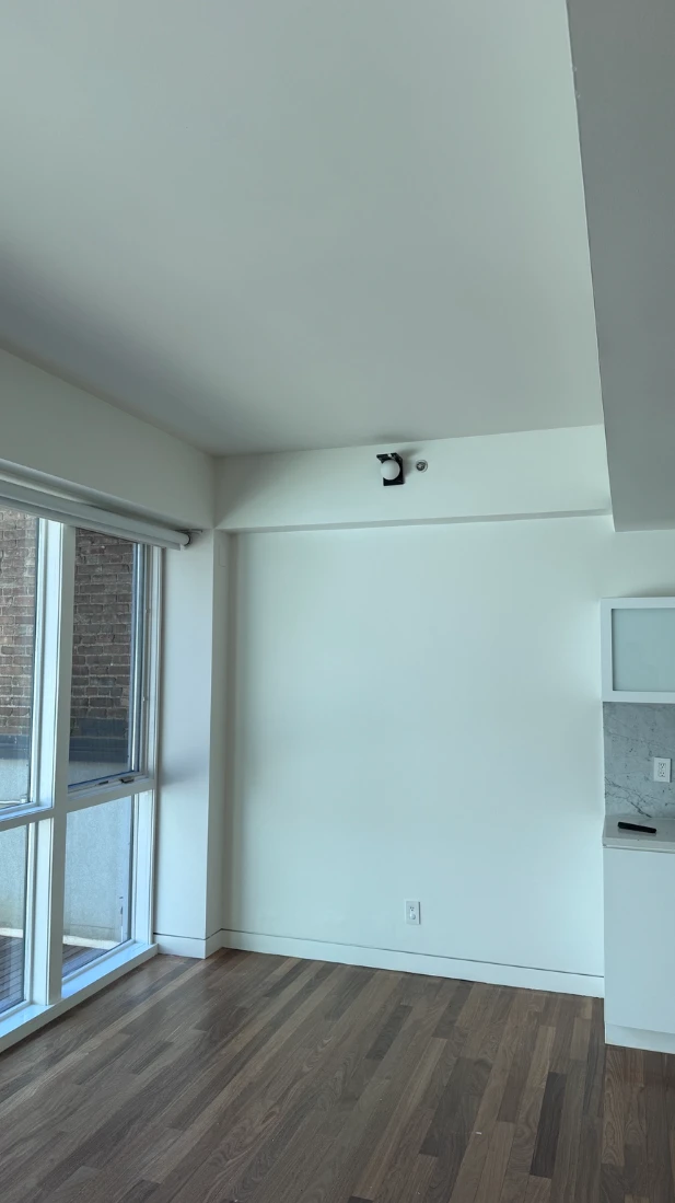 Corner of an apartment living area with smooth white walls and ceiling, a large window with a roller shade, white baseboards and hardwood floors