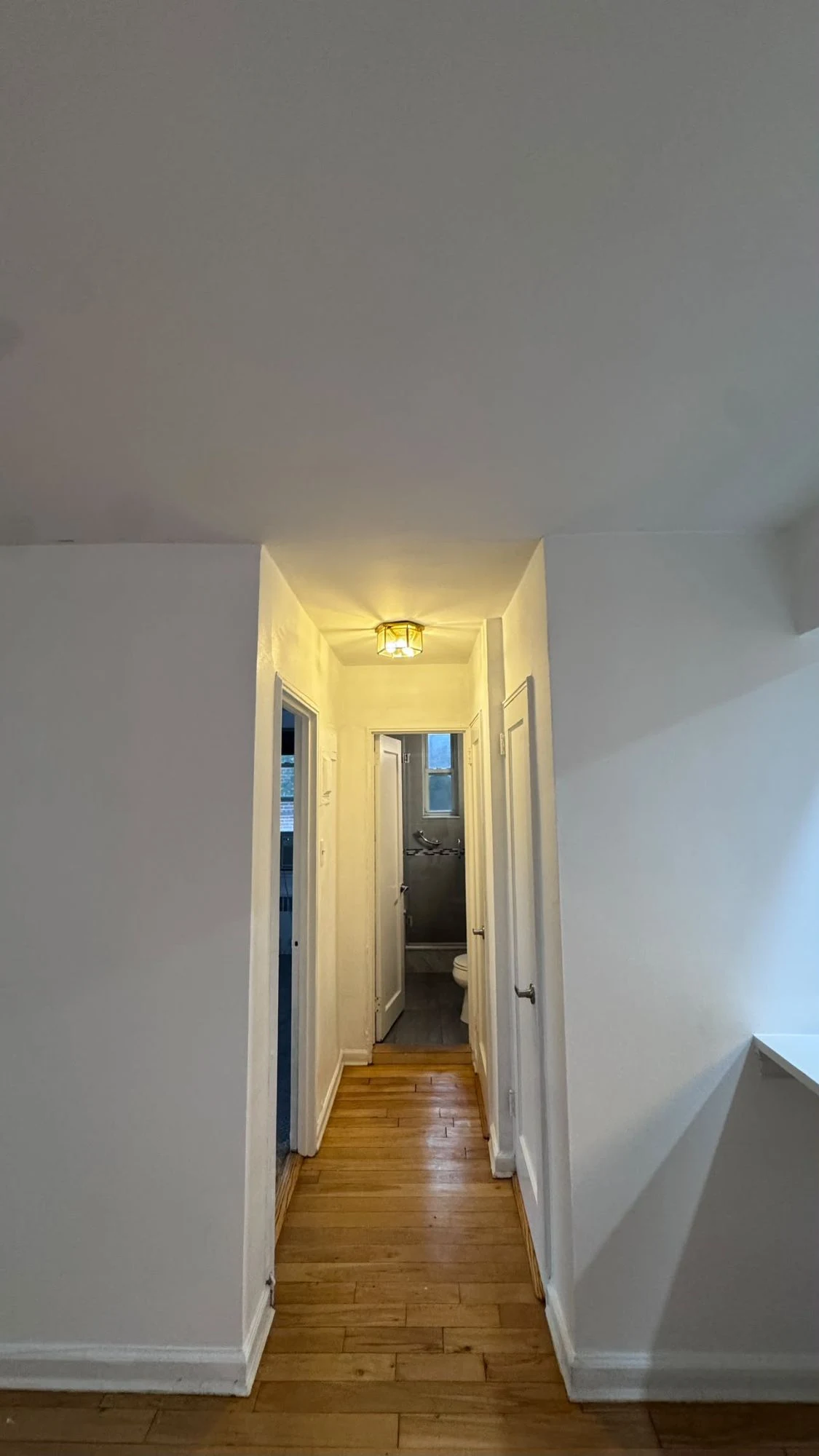 Apartment hallway with fresh white walls and ceiling, white doors and a view through to the bathroom, with hardwood floors