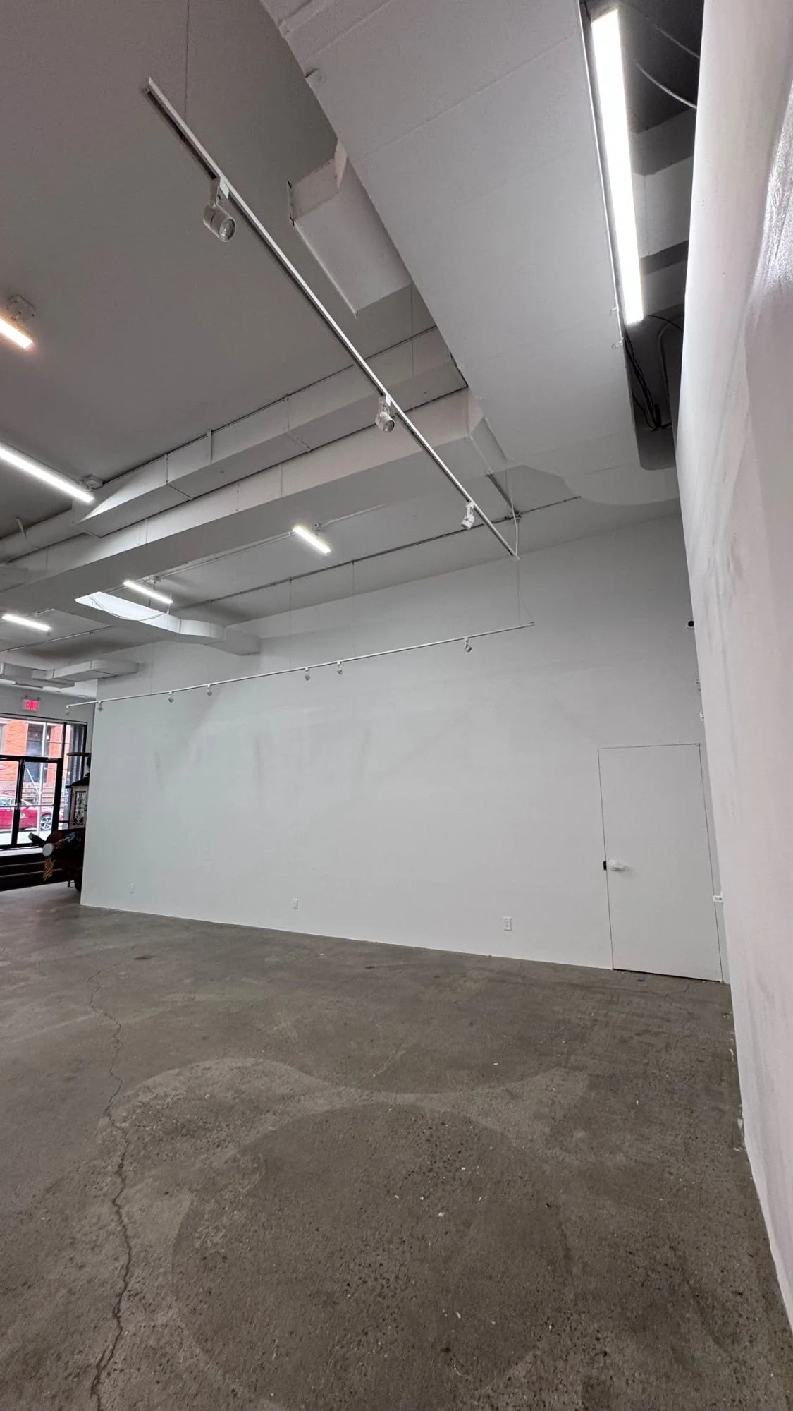 Tall, smooth white wall in a commercial space with track lighting, a white flush door, white ducts overhead and a concrete floor